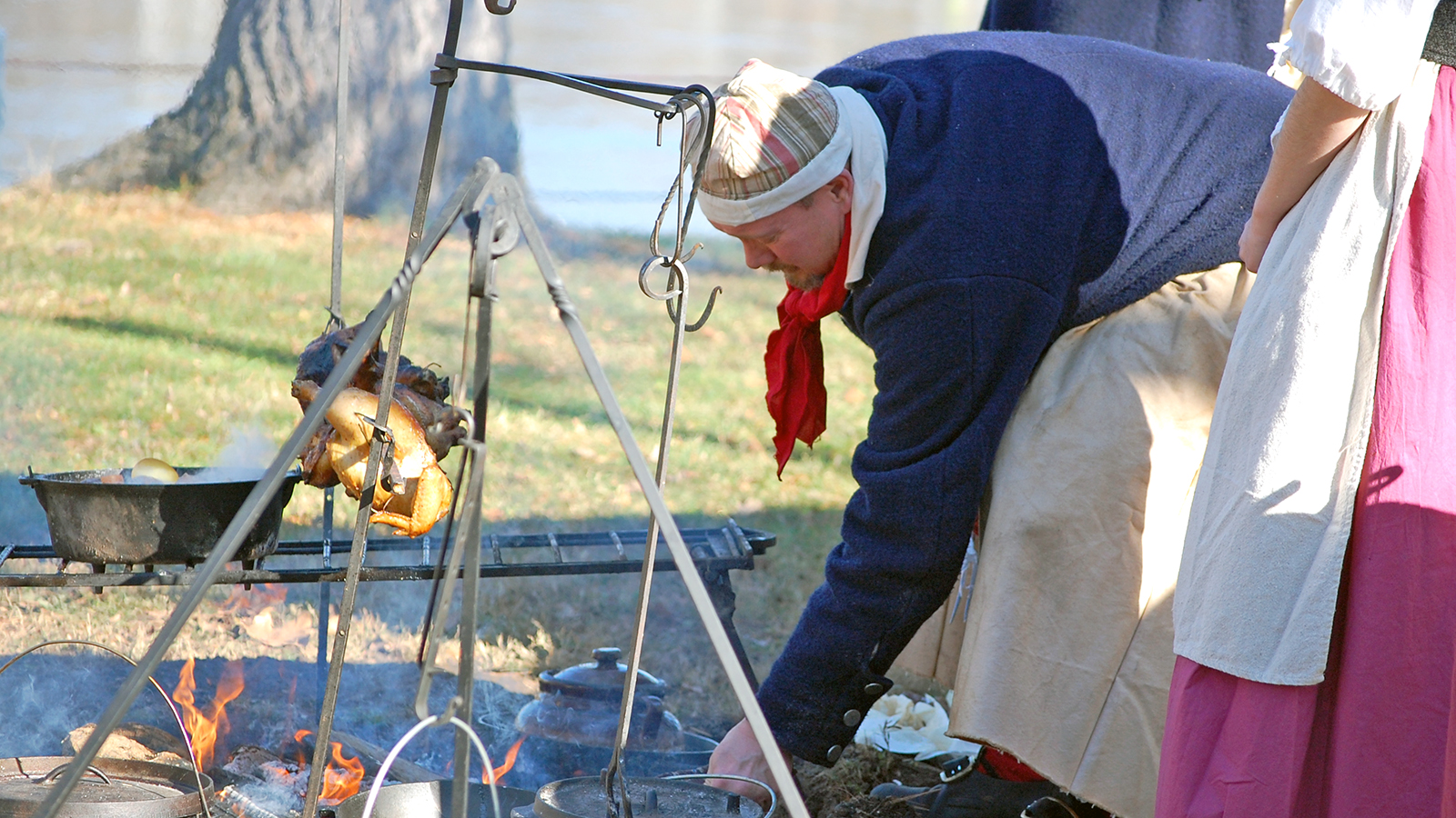 A man leans over a campfire being used to cook a chicken.
